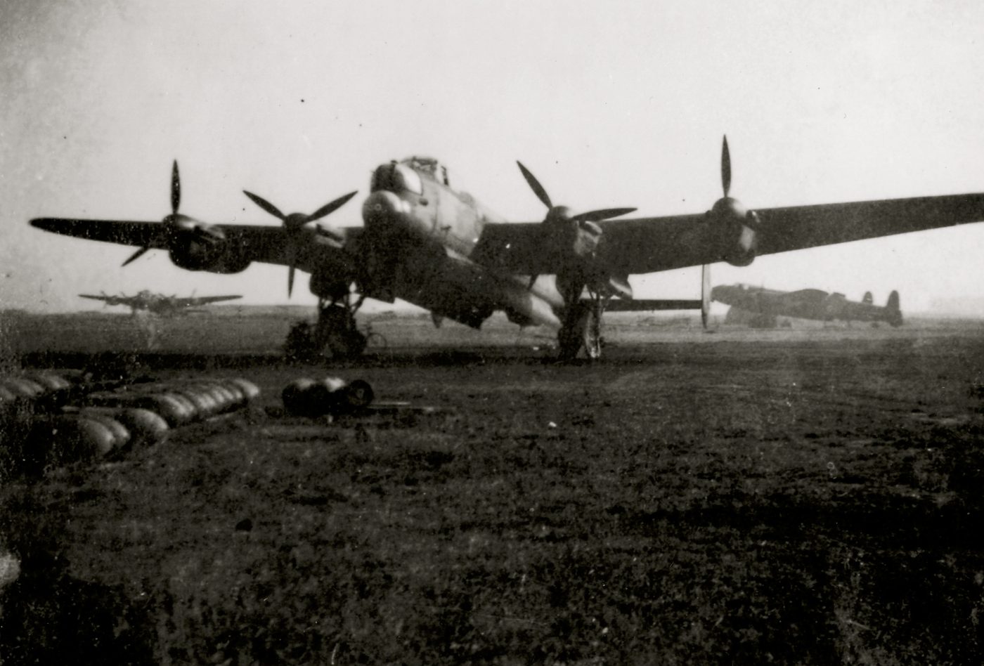 Lancaster bombers on the airfield with bombs laid out ready for loading, other aircraft visible in the misty background